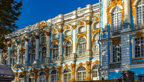 Фасад дворца с голубыми стенами, белыми колоннами и позолотой, освещённый солнцем на фоне синего неба