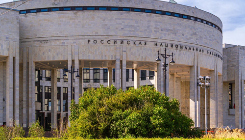 Фасад с колоннами и надписью «РОССИЙСКАЯ НАЦИОНАЛЬНАЯ БИБЛИОТЕКА»; спереди кустарник и уличные фонари