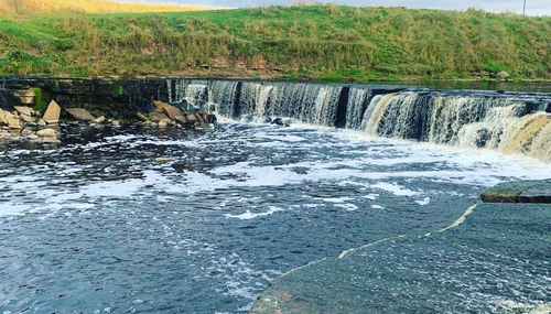 Низкий широкий водопад через каменный порог; бурная вода с пеной стекает в реку, на берегу травяной склон