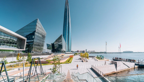 Высокая стеклянная башня у берега, рядом мост и набережная, отражение в спокойной воде, облачное небо