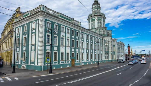 Кунсткамера: бирюзово-белое здание с башней на набережной; машины едут по дороге, знак пешеходного перехода