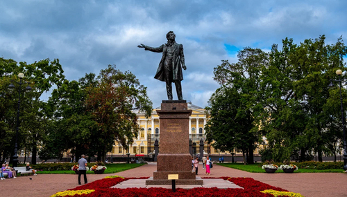Бронзовая статуя мужчины с вытянутой рукой на постаменте в парке; люди и цветники, сзади классическое здание