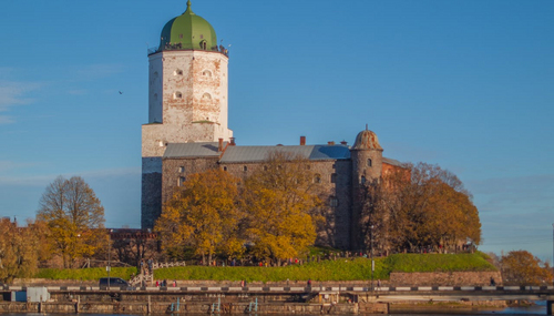 Каменная крепость с белой башней и зелёным куполом на берегу, осенние деревья и набережная в Выборге