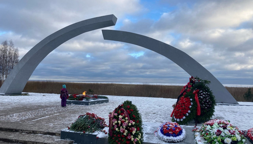 Две серые арки мемориала, у подножия вечный огонь и венки с цветами на снегу, человек стоит рядом у берега