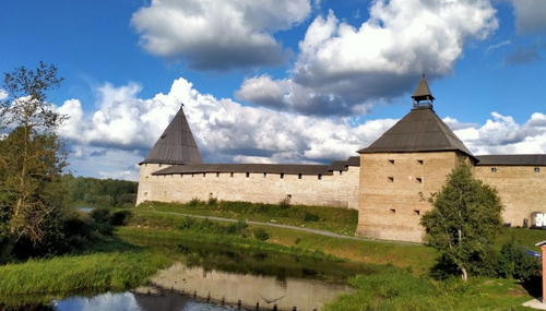 Каменные крепостные стены и две башни у воды, отражение в пруду, зелень и облачное летнее небо